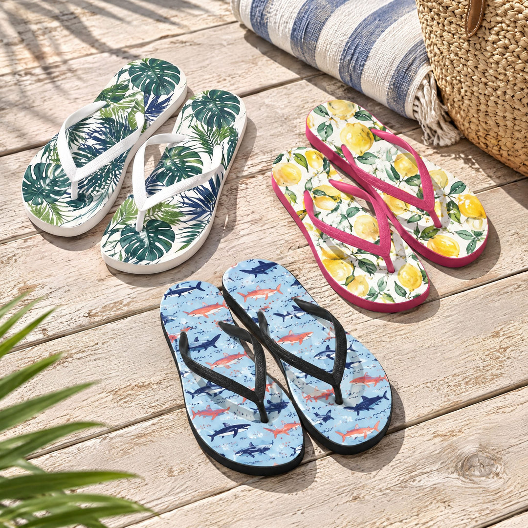 Adult sublimation flip flops: three printed pairs with tropical leaf, lemon and shark designs on a sunny wooden deck beside a striped towel and a wicker bag