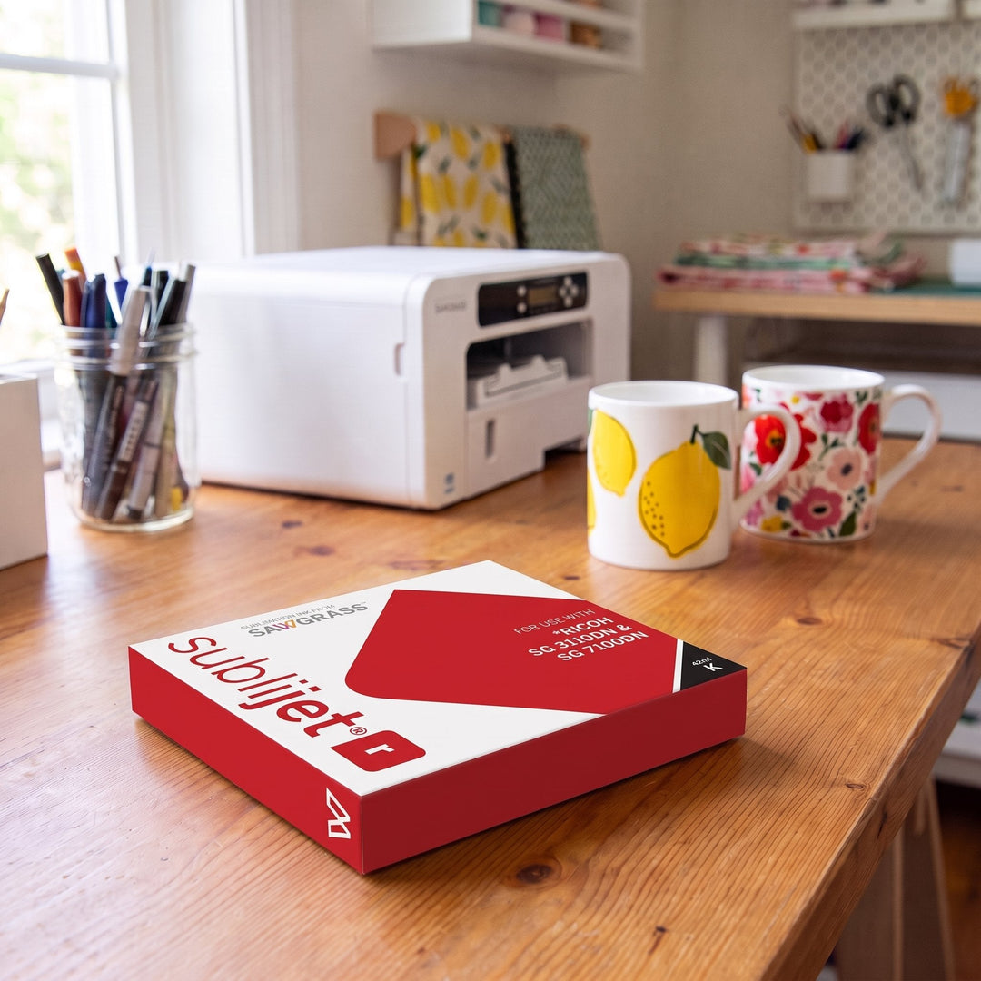 Sublijet-R ink for Ricoh SG7100DN: a red SubliJet R cartridge box on a wooden desk with lemon and floral print mugs and a white printer behind