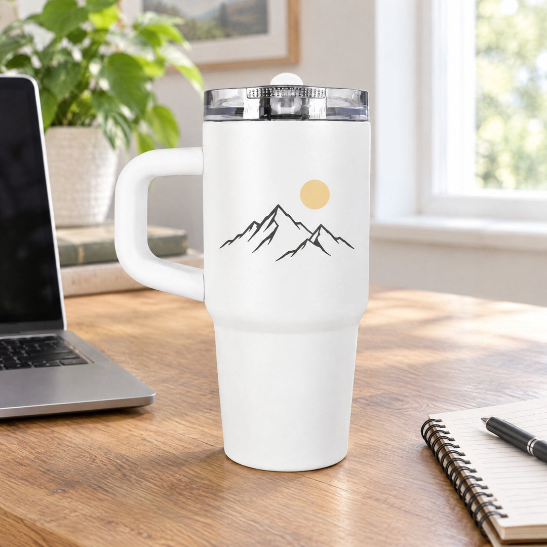 14oz sublimation tumbler printed with line-art mountains and a yellow sun, on a wooden desk beside a laptop
